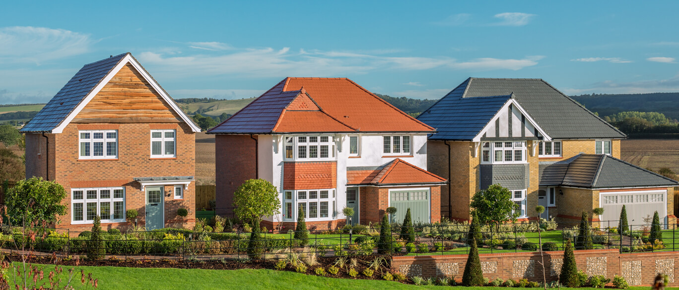 A row of modern detached houses with colourful roofs and well-maintained gardens, set against a rural landscape under a blue sky.