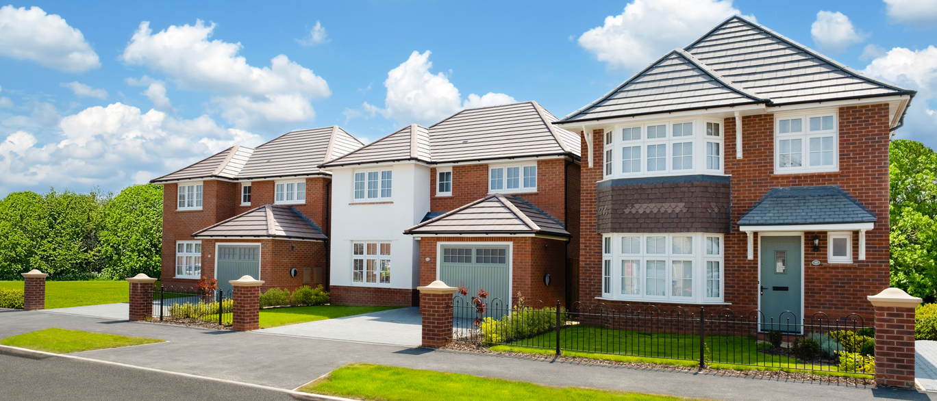 A row of modern red-brick houses with grey tiled roofs under a blue sky with fluffy clouds, surrounded by green lawns and trees at The Hollies at Great Milton Park
