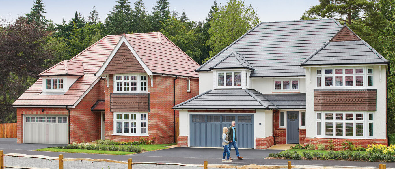 Two people walking in front of a modern semi-detached house with a red brick and white exterior, surrounded by greenery at Hendricks Green.