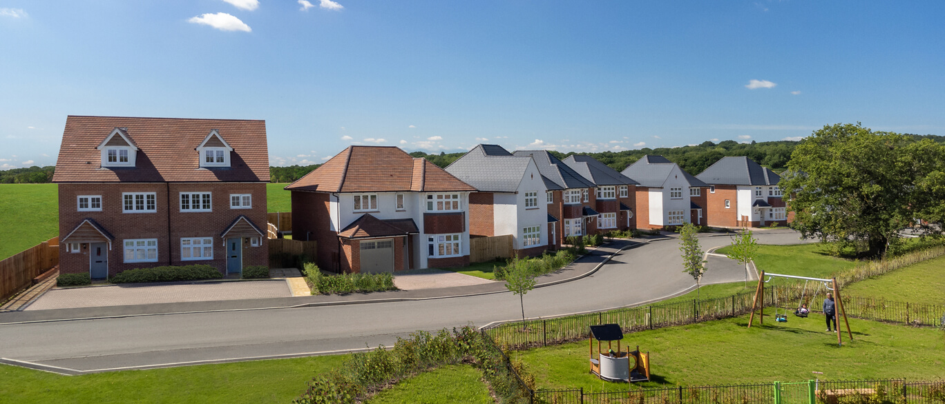 Newly built suburban houses with front gardens, a curved road, and a grassy area with a swing set under a blue sky at Westley Green.