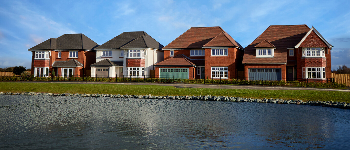 Modern suburban houses overlooking a lake with a paved pathway and well-kept gardens under a blue sky at Curborough Lakes.