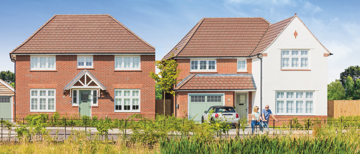 Two modern detached houses with red brick and white facades, front gardens, and a couple walking along the pavement on a clear day at Hugglescote Grange.