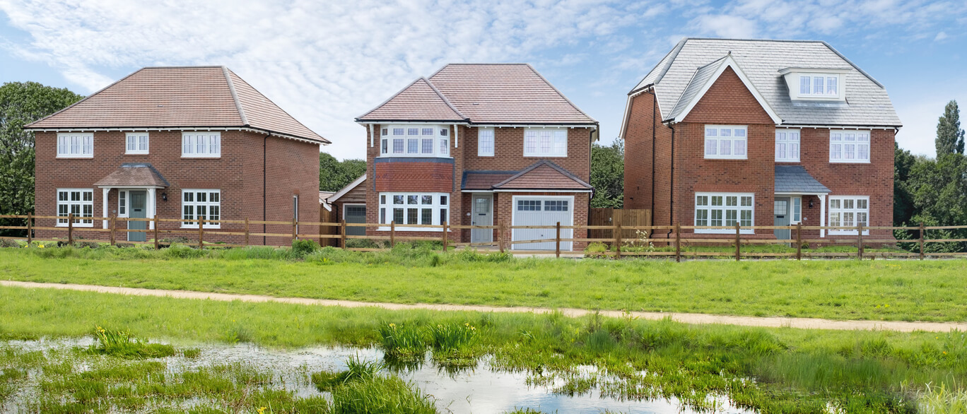 Three newly built red-brick houses with grey and brown tiled roofs, large white-framed windows, set behind a wooden fence with a grassy field in the foreground at Midsummer Meadow.