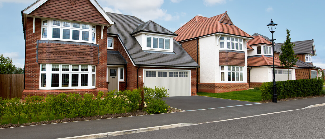 Modern detached houses with bay windows, red and white exteriors, and well-maintained front gardens on a suburban street in Britain at Mill Green Meadows.
