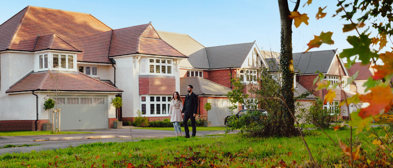 Suburban street with large two-story houses, a couple walking on the sidewalk, and autumn leaves on the grass under a clear blue sky at Paddock Green.