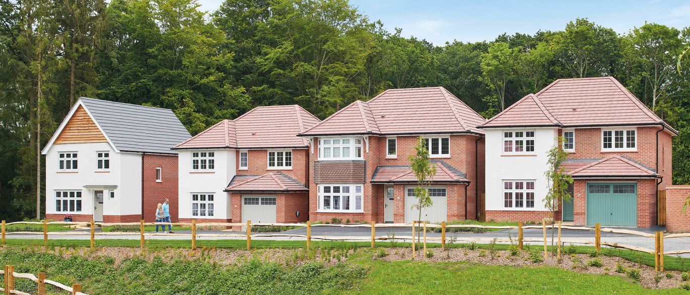 Newly built houses with red brick and white exteriors, some with bay windows, set against a green wooded background in a residential development.