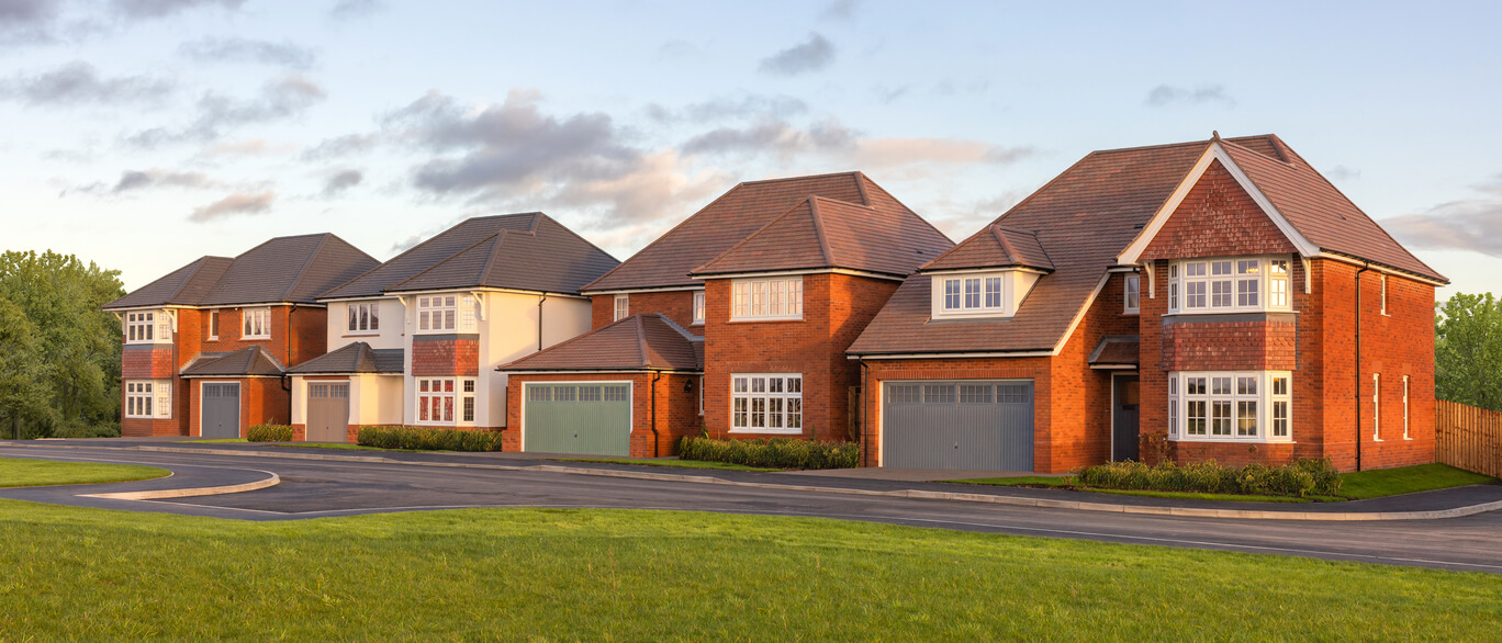 A row of modern red-brick and white houses with pitched roofs, set against a partly cloudy sky and surrounded by green grass and trees.