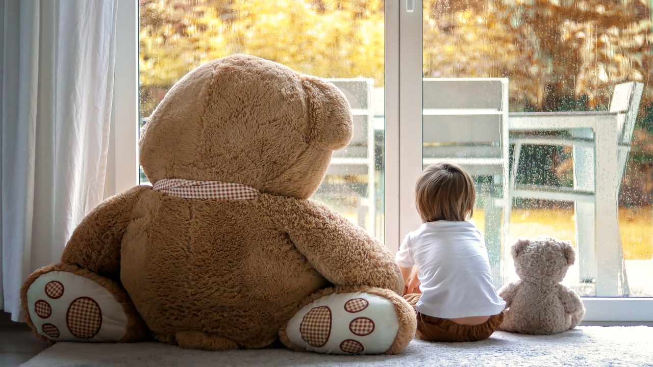 Small child with two teddy bears looking out of the window into the garden