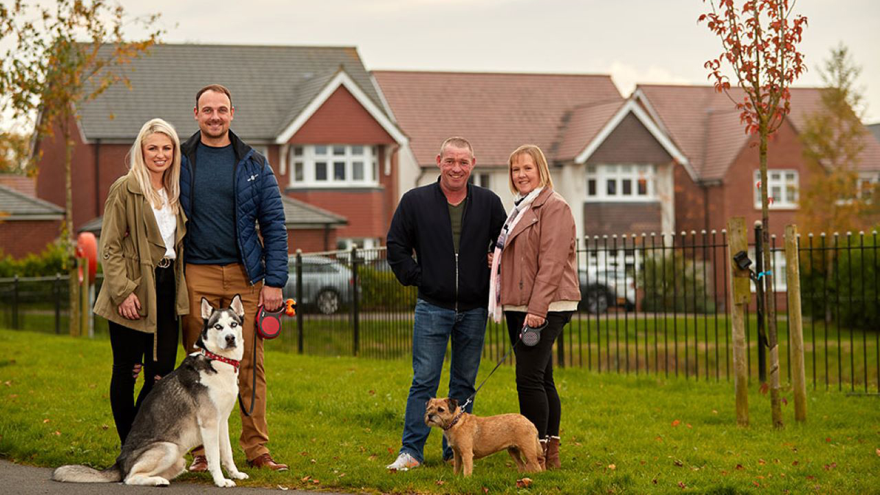 Two couples with dogs standing on the grass opposite a row of Redrow homes