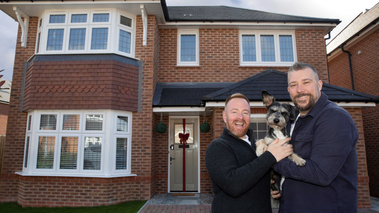 Couple cuddling their small dog outside their new home, which has a red bow tied around the front door
