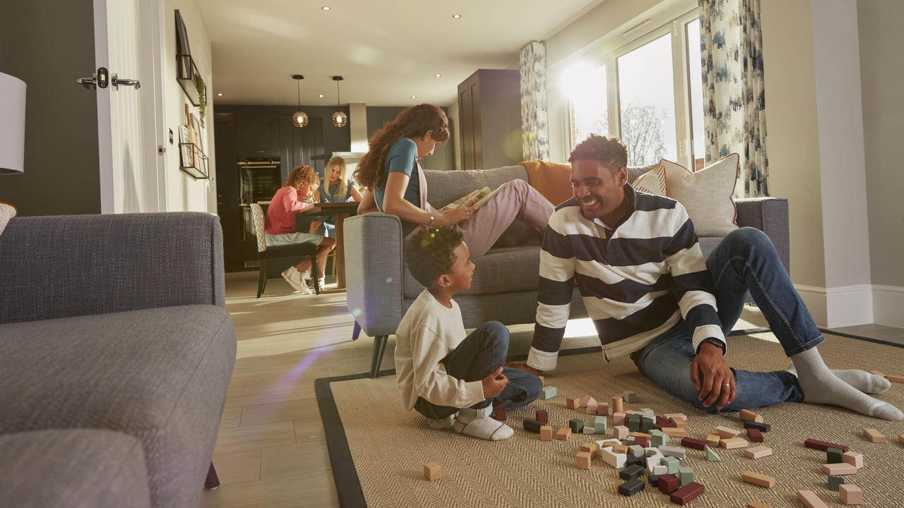 A father and son playing with blocks on the floor while an older child reads on the sofa