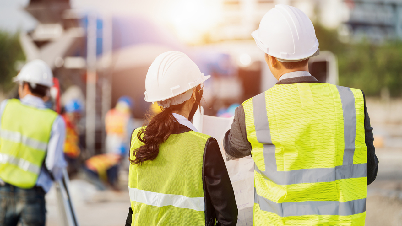 Two construction workers in hard hats and hi vis vests looking at a large sheet of paper