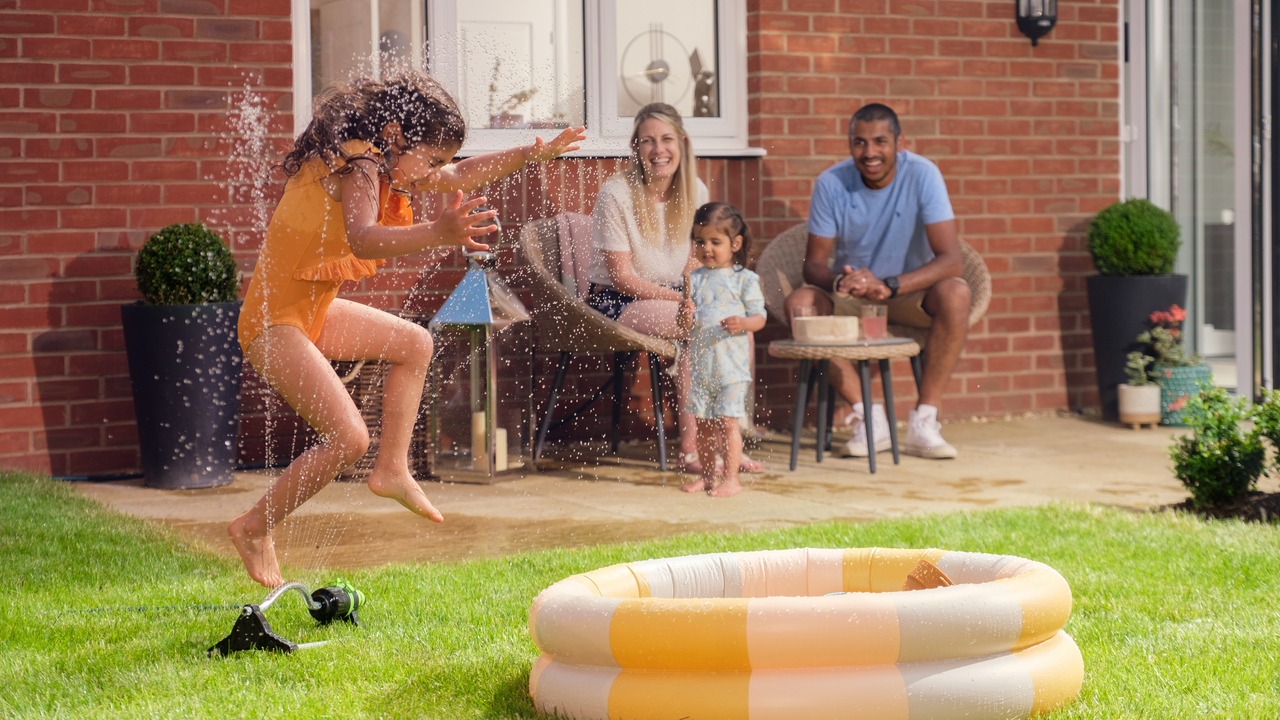 Little girl jumping over a water sprinkler while smiling family watches on