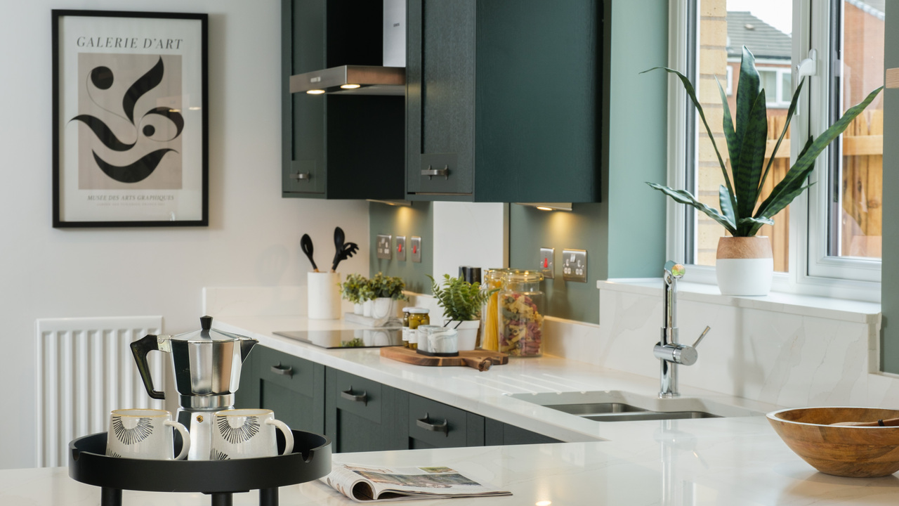 A sink by a window in a white kitchen with dark green cupboards