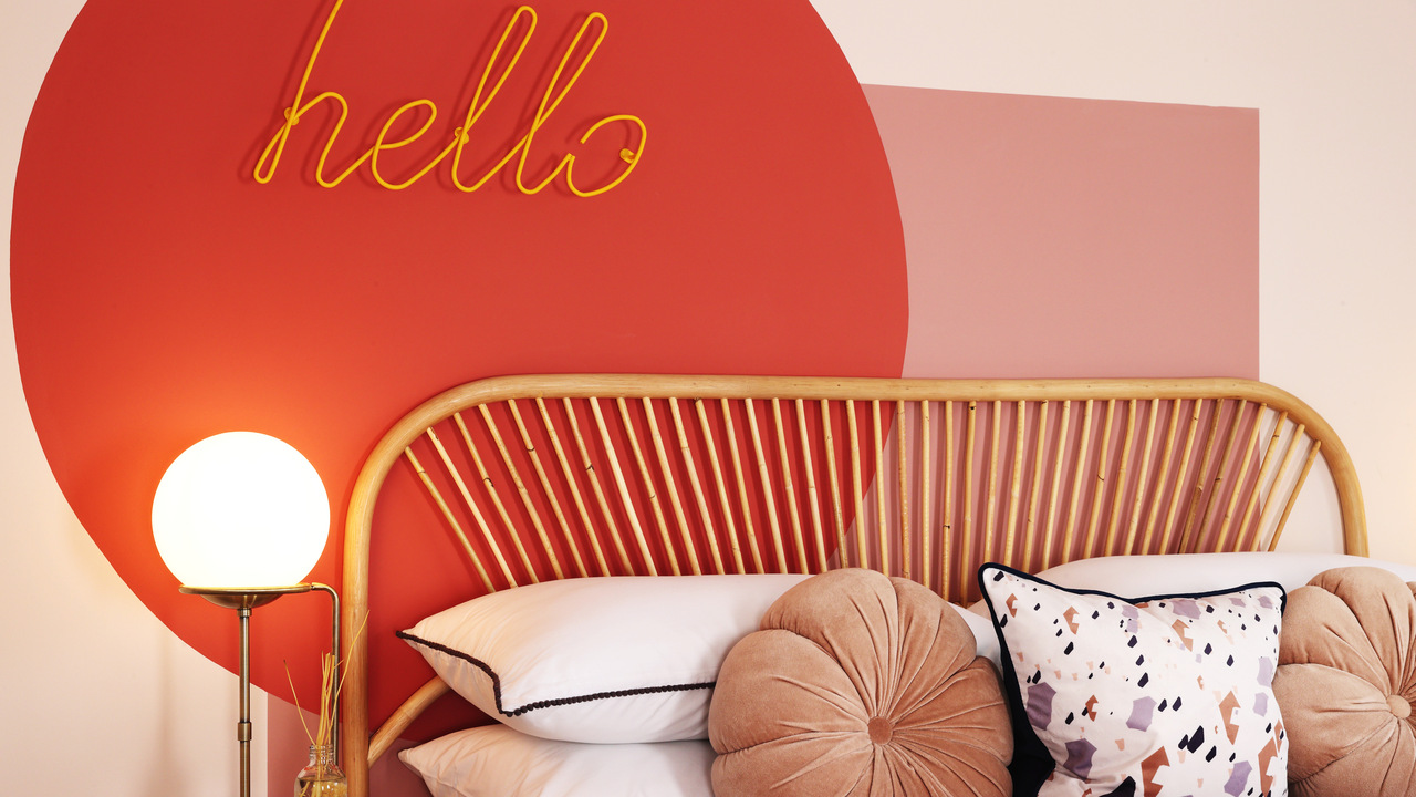 Neon sign saying "hello" hung on a wall painted with red and pink geometric shapes over a bed with a rattan frame
