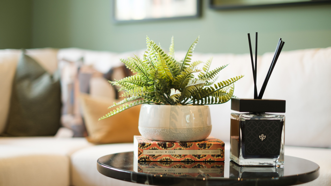 White corner sofa and a small, black side table featuring a Redrow reed diffuser and a potted plant