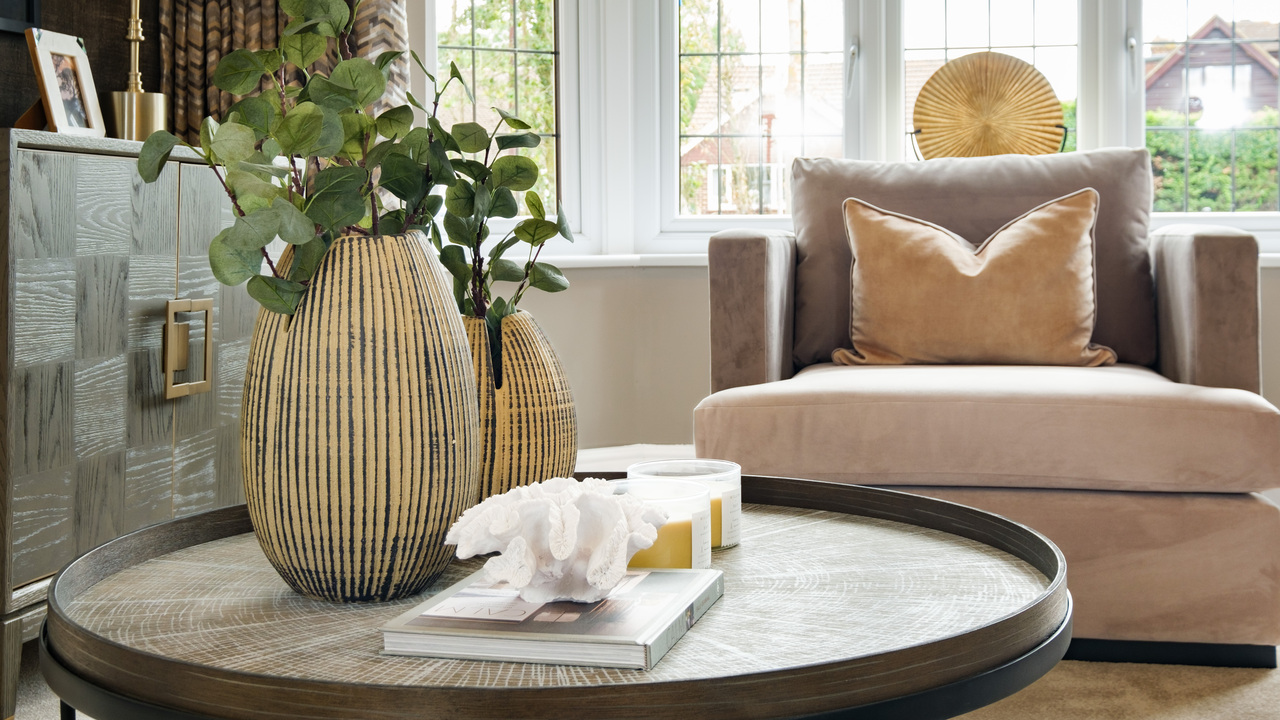 Low wooden side table featuring potted plants, a shell and a candle. A beige chair sits in a bay window.