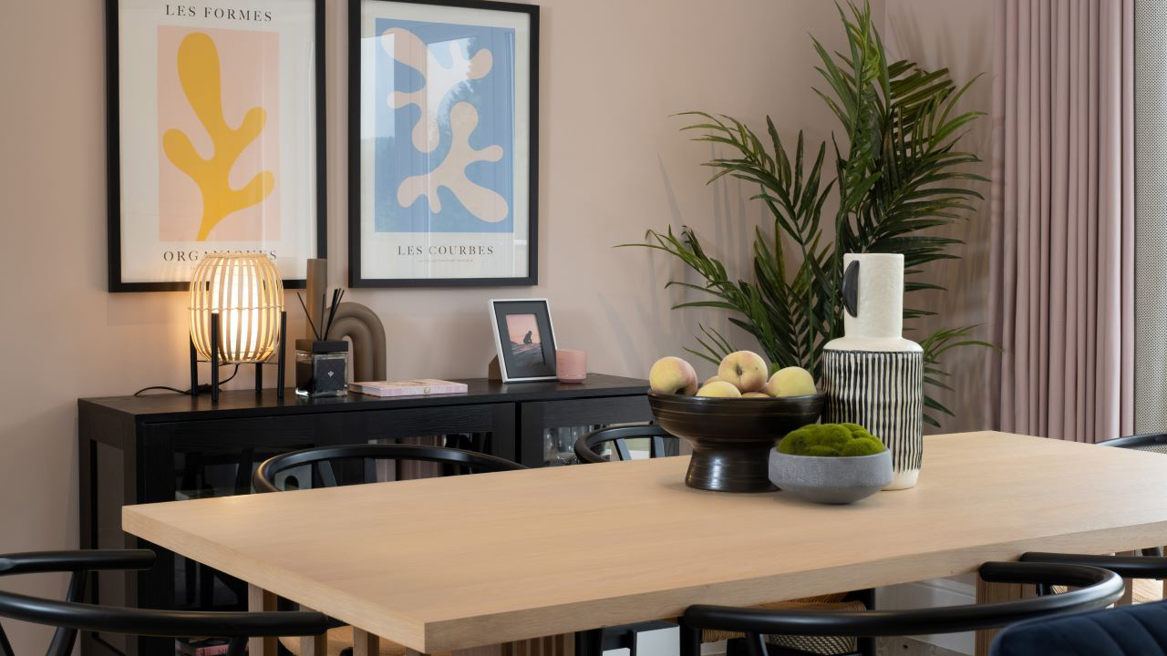 Modern, wood dining table with black chairs. The top of the table features a bowl of fruit, a large vase and a bowl of moss.