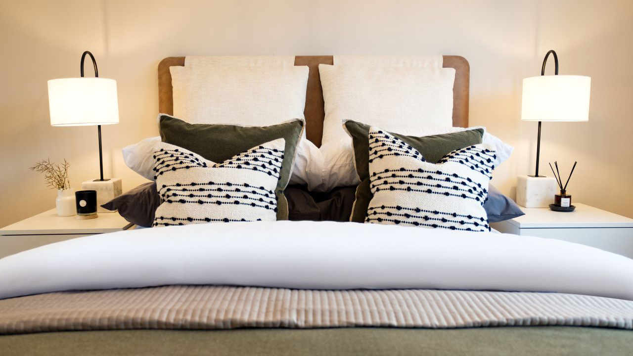 Bed with multiple rows of textured cushions. There is a bedside table on each side with matching lamps.