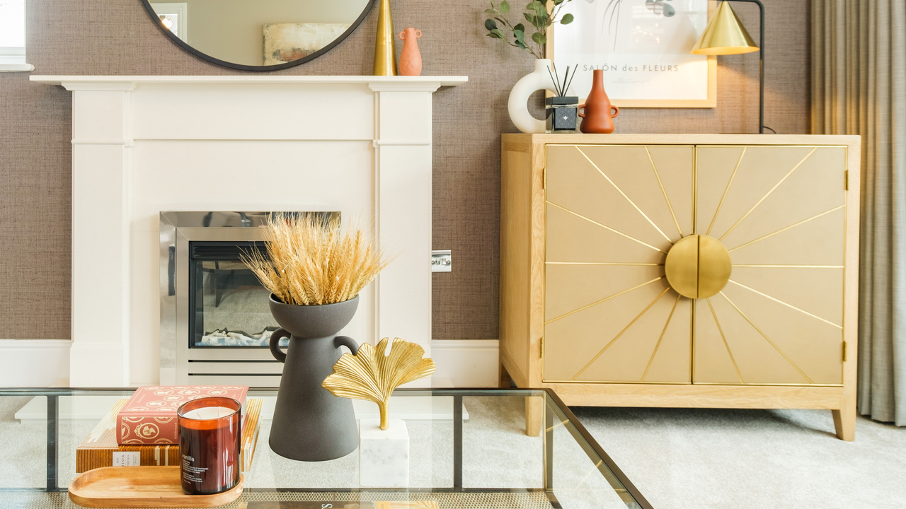Corner of a lounge featuring a white fireplace, glass coffee table and a cabinet with gold details that look like a sun