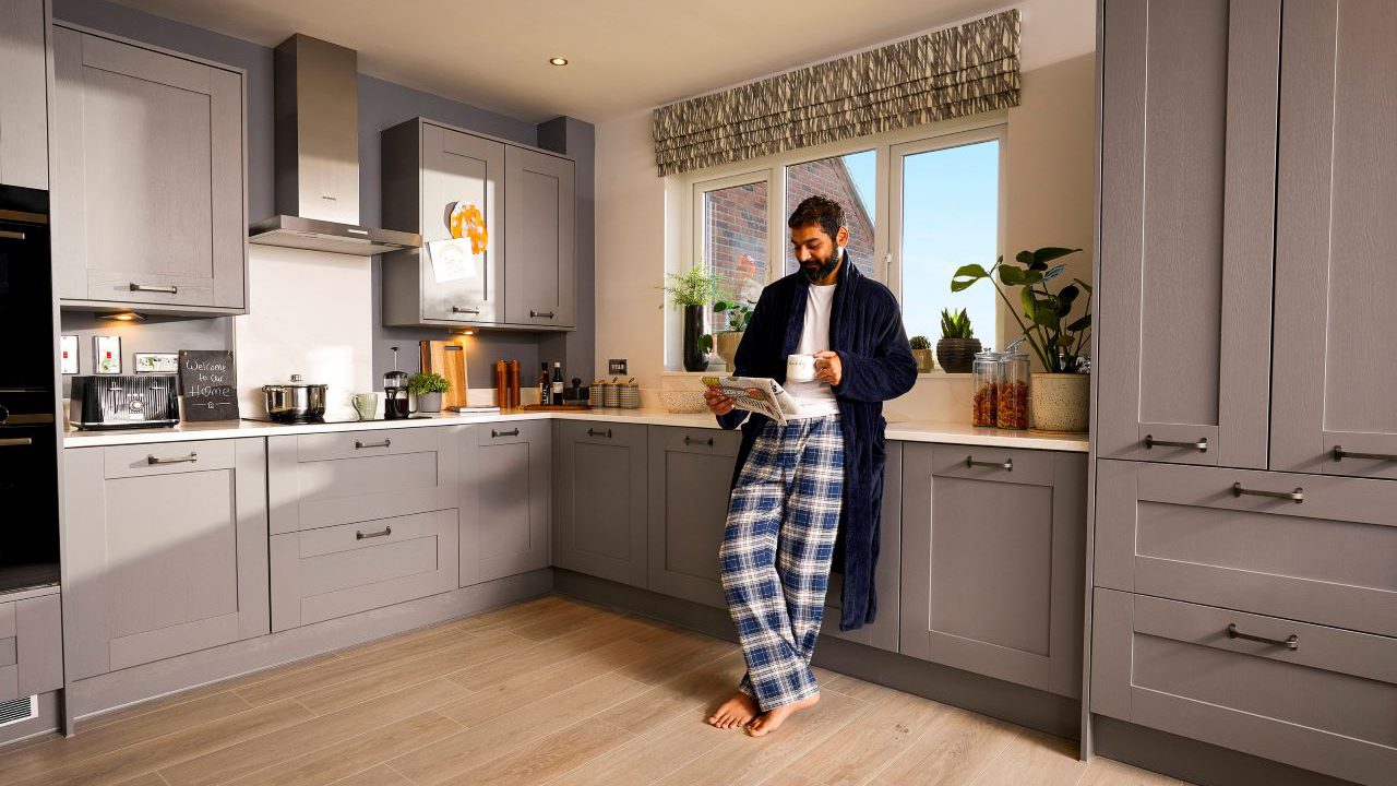 Man in a dressing gown reading a newspaper while leaning against his kitchen counter