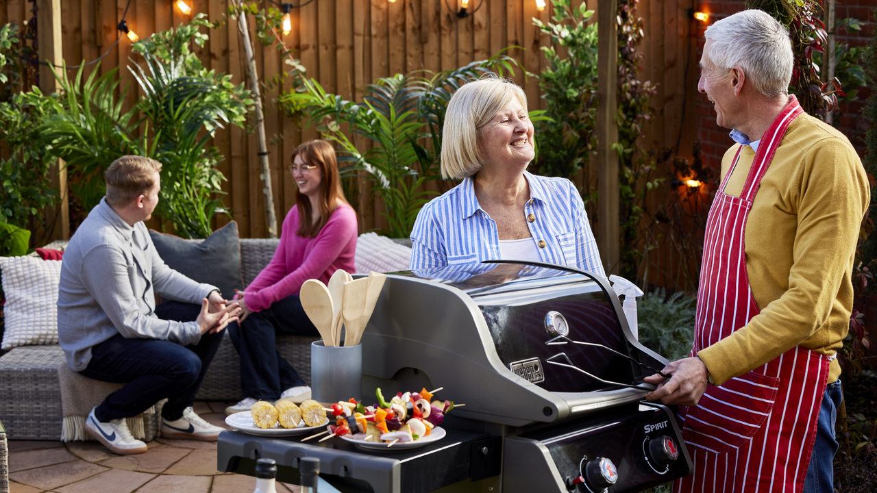 One couple chatting next to a barbeque while another sits on outdoor seating in the background