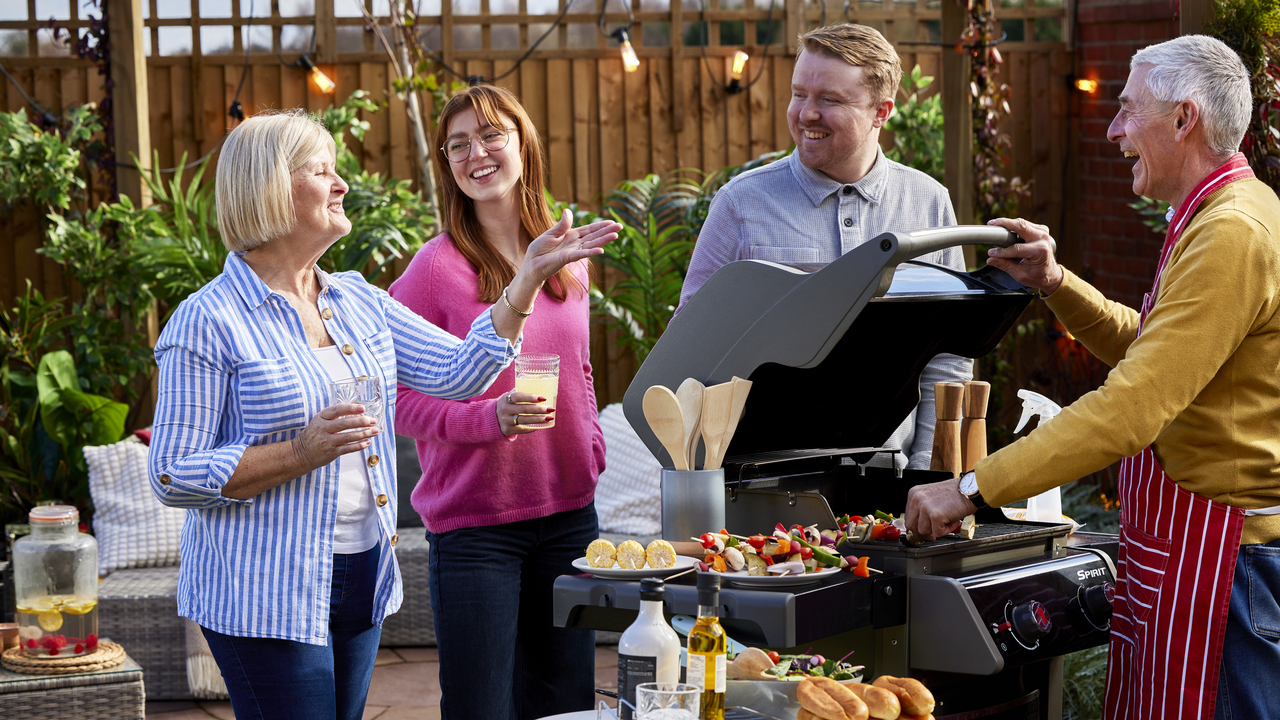 Four people having a chat in the garden as a man places vegetable skewers on a barbeque 