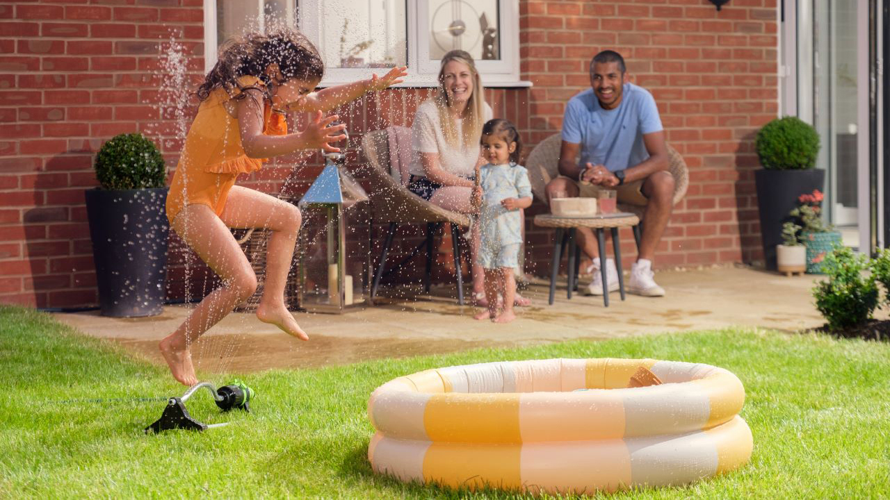 Family sitting in their garden, smiling as a little girl jumps through a water sprinkler