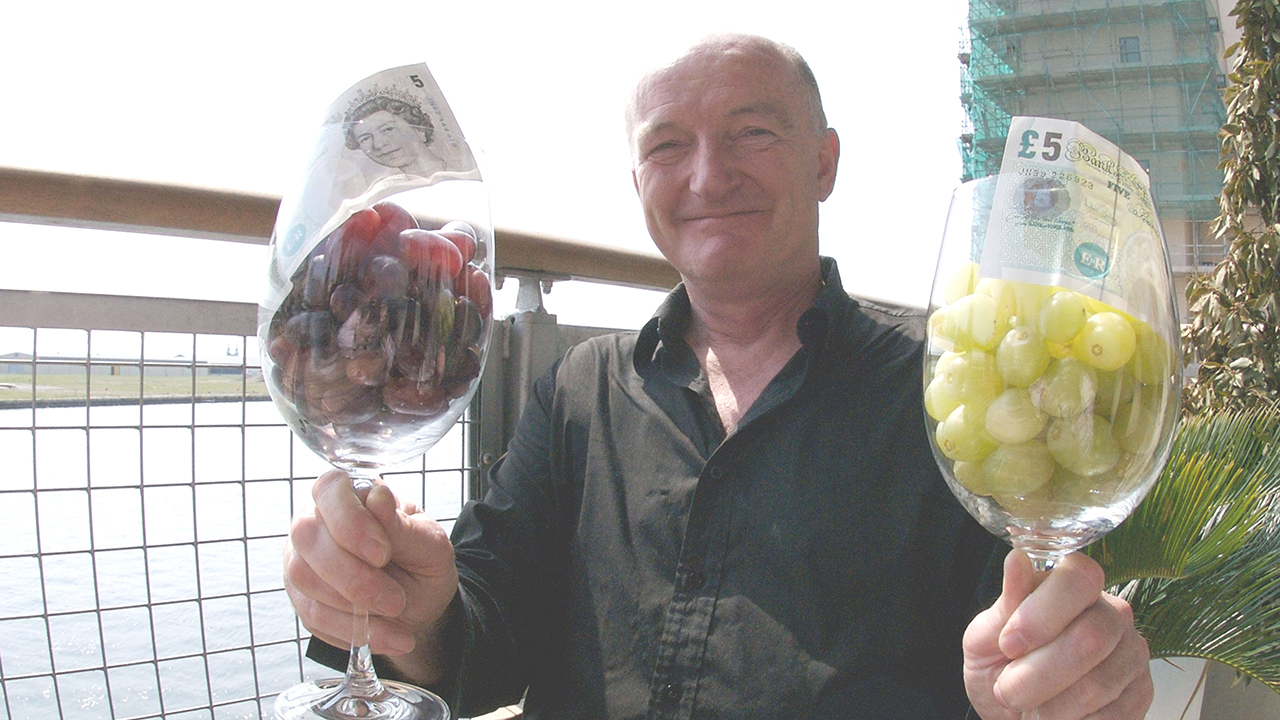Oz Clarke holding two large wine glasses containing grapes and £5 notes