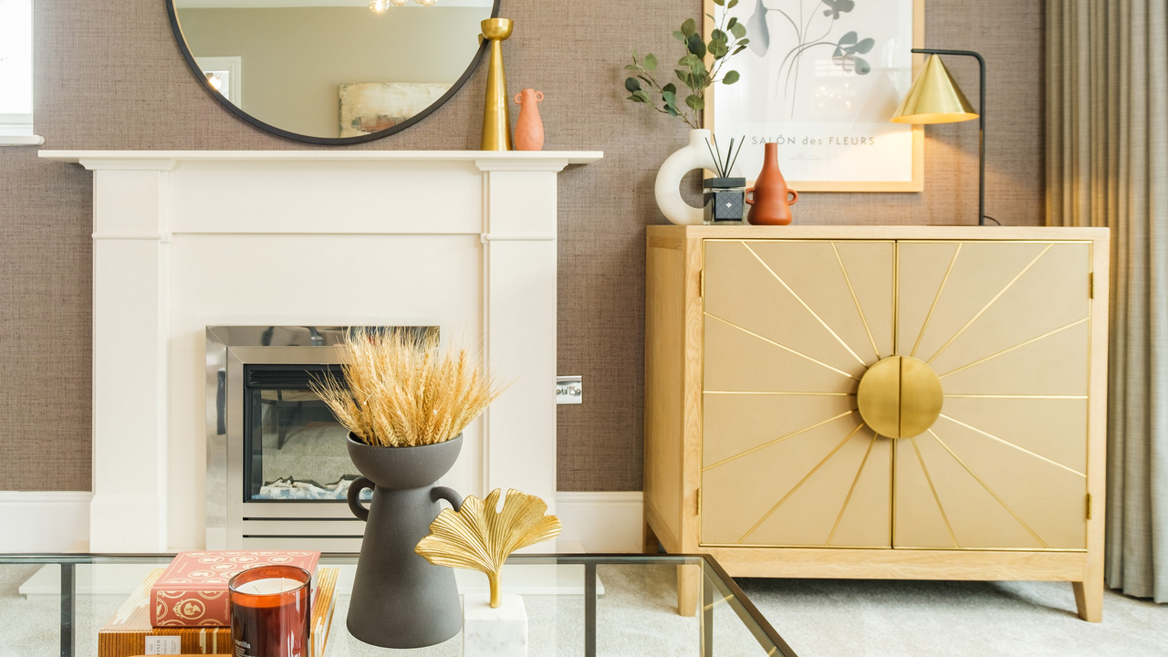 Area of a lounge showing a glass coffee table, white fireplace and cupboard with gold details