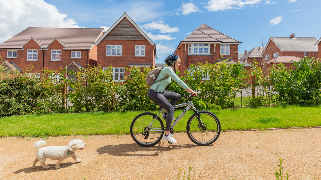 Woman cycling down a path by a Redrow development with a little white dog running alongside her